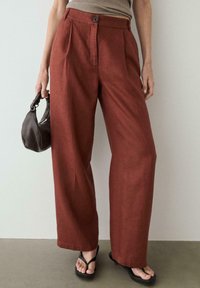 Femme portant un pantalon taille haute à jambes larges couleur rouille, un haut beige, des sandales noires fines et tenant un petit sac à main noir.
