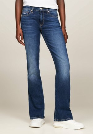 Jeans de mezclilla azules acampanados con una cintura de media altura, que presentan un diseño clásico de cinco bolsillos y un ligero desgastado en los muslos, combinados con zapatillas blancas.