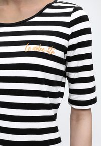 Chemise à manches courtes rayée noir et blanc, avec le texte brodé en orange "la dolce vita" sur le côté gauche de la poitrine.