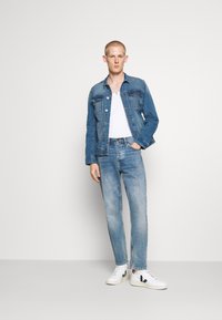 Jeansjacka, ljusblå, knäppt framtill med två bröstfickor; vit t-shirt under; ljusblå jeans; vita sneakers med svart logotyp.