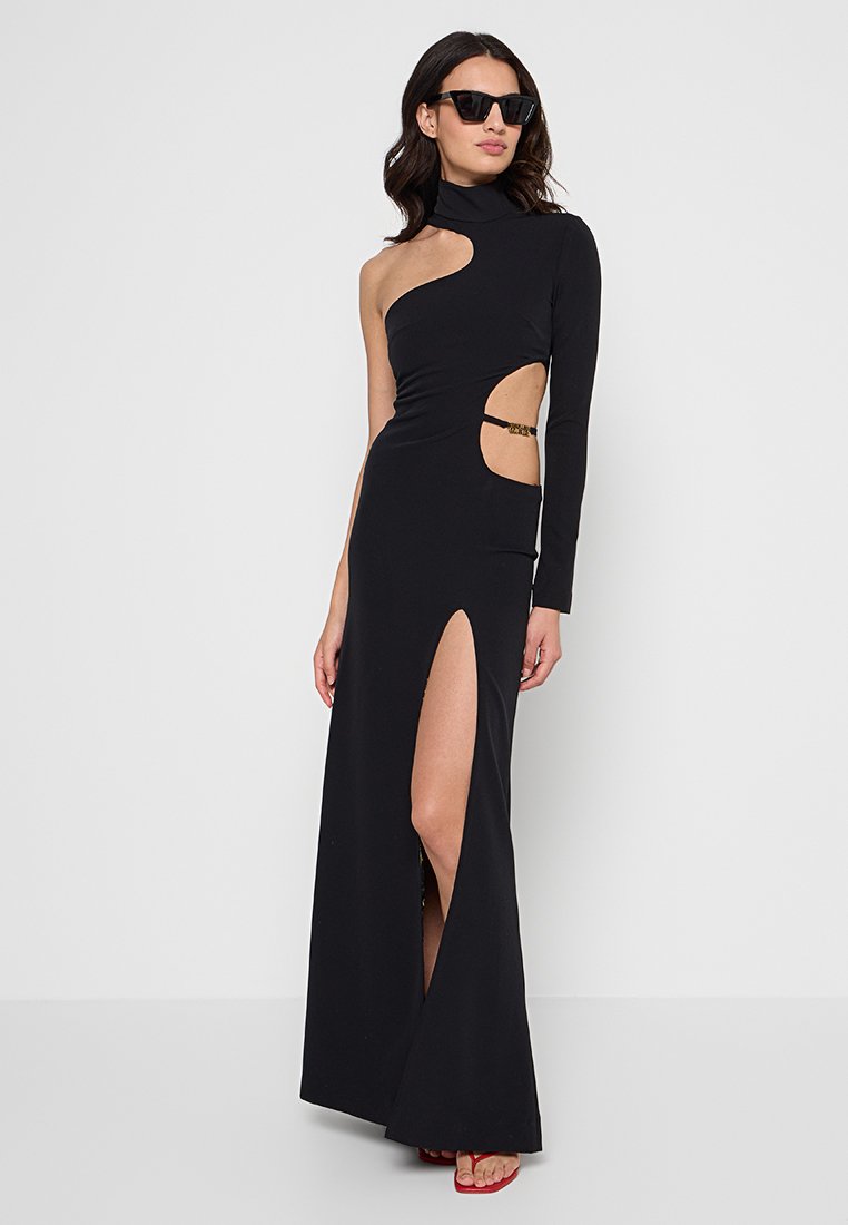 Femme portant une robe noire asymétrique avec découpes, fente haute, manches longues, col roulé et lunettes de soleil noires sur fond uni.