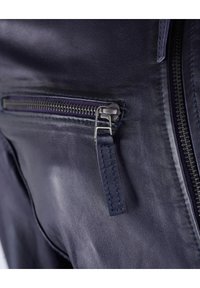 Nahaufnahme einer navyblauen Lederjacke, die eine silberne Reißverschlusstasche mit strukturiertem Oberflächen und genähten Akzenten zeigt, wodurch das Material und das Design hervorgehoben werden.