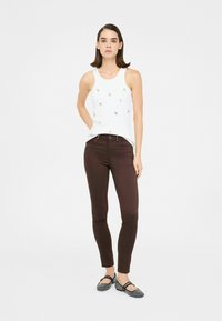 Canotta bianca con piccole decorazioni, pantaloni aderenti marroni e flats neri, su un modello contro uno sfondo neutro.