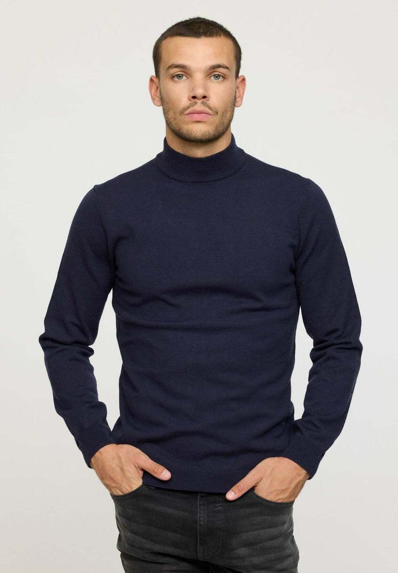 Pull à col roulé bleu marine en tissu tricoté, design ajusté avec des manches longues, col et ourlet côtelés, porté avec les mains devant.