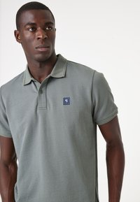 Polohemd in hellgrau mit einem strukturierten Muster, dreiknöpfiger Blende und einem kleinen blauen Logo auf der linken Brust. Kurze Ärmel.