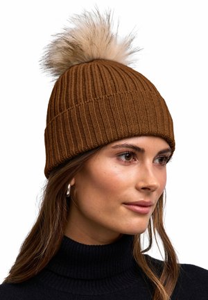 Kobieta nosząca brązową prążkowaną dzianą czapkę z dużym beżowo-brązowym pomponem ze sztucznego futra, czarny golf i srebrne kolczyki koła.