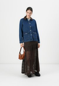 Jeansjakke med brun faux fur-krage, to frontlommer, båret over et langt brunt blonderkjørt. Modellen holder en brun veske og står foran en ensfarget bakgrunn.