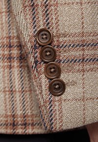 Beżowa i brązowa tkanina w wzór houndstooth z czterema brązowymi guzikami na rękawie kurtki, ukazująca teksturowany splot i kontrastujące szwy.