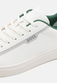 Valge nahast sneaker, millel on rohelised nüansid, tekstureeritud kummist talla ja hall "BOSS" logo küljel. Nöörid on valged ja lamedad.