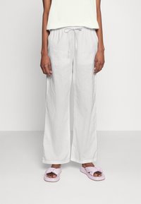 Femme portant un pantalon en lin blanc ample avec taille élastique et des sandales légères en mousse rose clair, se tenant devant un fond uni.
