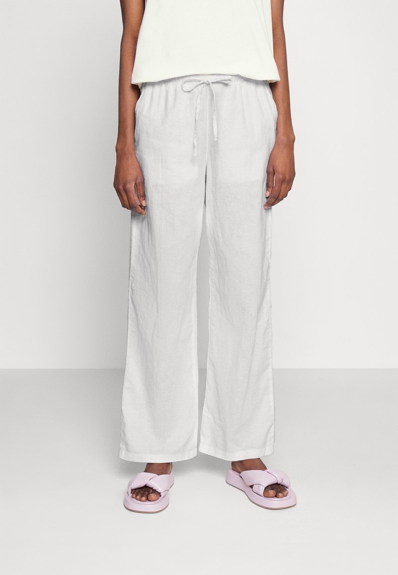 Femme portant un pantalon en lin blanc ample avec taille élastique et des sandales légères en mousse rose clair, se tenant devant un fond uni.