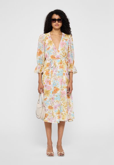 Floral midi dress with a V-neckline, three-quarter puff sleeves, pastel hues, and an adjustable waist tie. Accompanied by a small handbag.