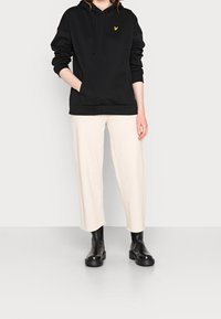 Sweatshirt à capuche noir avec une poche avant et un logo jaune, associé à un pantalon crème à jambes larges et des bottes noires chunky à la cheville.