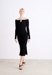 Black ribbed knit dress with long sleeves, off-the-shoulder design, keyhole detail, and side slit, paired with multicolored high-heeled sandals.