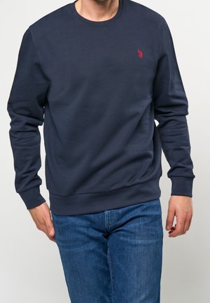 Mann trägt marineblauen Pullover mit langen Ärmeln und kleinem roten Logo auf der Brust sowie blaue Jeans, steht vor einem schlichten, hellen Hintergrund.