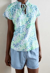Blouse à manches courtes en tissu léger avec un motif floral bleu et vert, dotée d'une fermeture à nouer au niveau du col et de détails froncés.