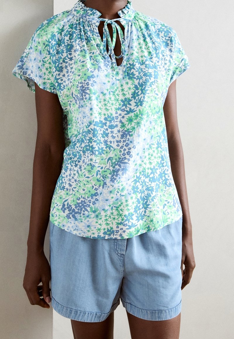 Blouse à manches courtes en tissu léger avec un motif floral bleu et vert, dotée d'une fermeture à nouer au niveau du col et de détails froncés.
