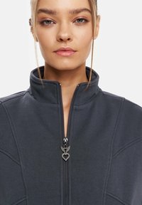 Dunkelgraue Zip-Jacke mit hohem Kragen, die strukturierte Nähte und einen herzförmigen Reißverschlussanzug hat. Weicher Stoff mit lässigem Design.