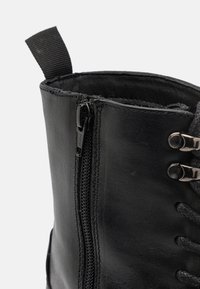Botín negro de cuero hasta el tobillo con cremallera lateral, cordones en la parte frontal y un acabado texturizado. En la parte superior hay una tira de tela para tirar.
