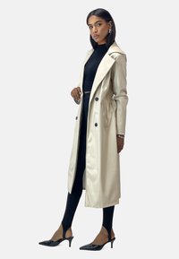 Lange, lichte beige trenchcoat met een matte afwerking, grote revers, dubbele knopen en zijzakken. Gecombineerd met een zwart aansluitend topje en smalle broek.
