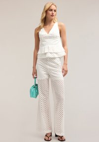 Witte oogjes jumpsuit met een peplum taille, v-hals en wijde pijpen, voorzien van uitsnijdingen en gecombineerd met een teal handtas.