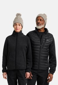 Giacca in pile nera con colletto alto, abbinata a un gilet imbottito nero. Entrambi indossano berretti lavorati a maglia grigi chiari con pon-pon.