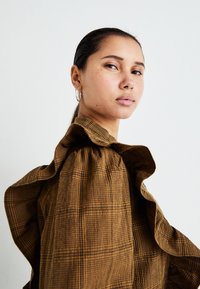 Blusa in tessuto a quadri marrone con colletto arricciato, caratterizzata da una finitura testurizzata e una vestibilità rilassata. Il modello ha una coda di cavallo liscia e orecchini a cerchio.