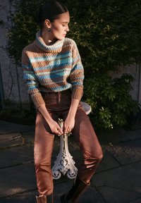 Strickpullover mit einem Chevron-Muster in Blau, Braun und Beige, kombiniert mit glänzenden braunen Hosen und schwarzen Stiefeletten.