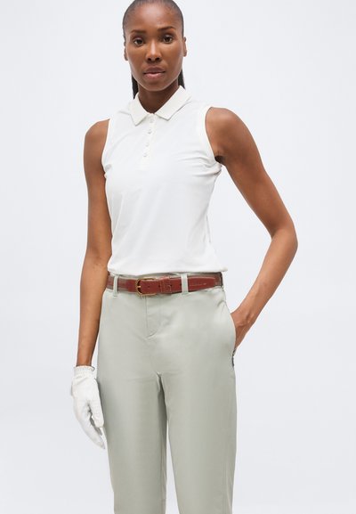 Femme portant un polo blanc sans manches, un pantalon gris clair avec une ceinture marron et un gant de golf blanc sur la main gauche, debout devant un fond uni.