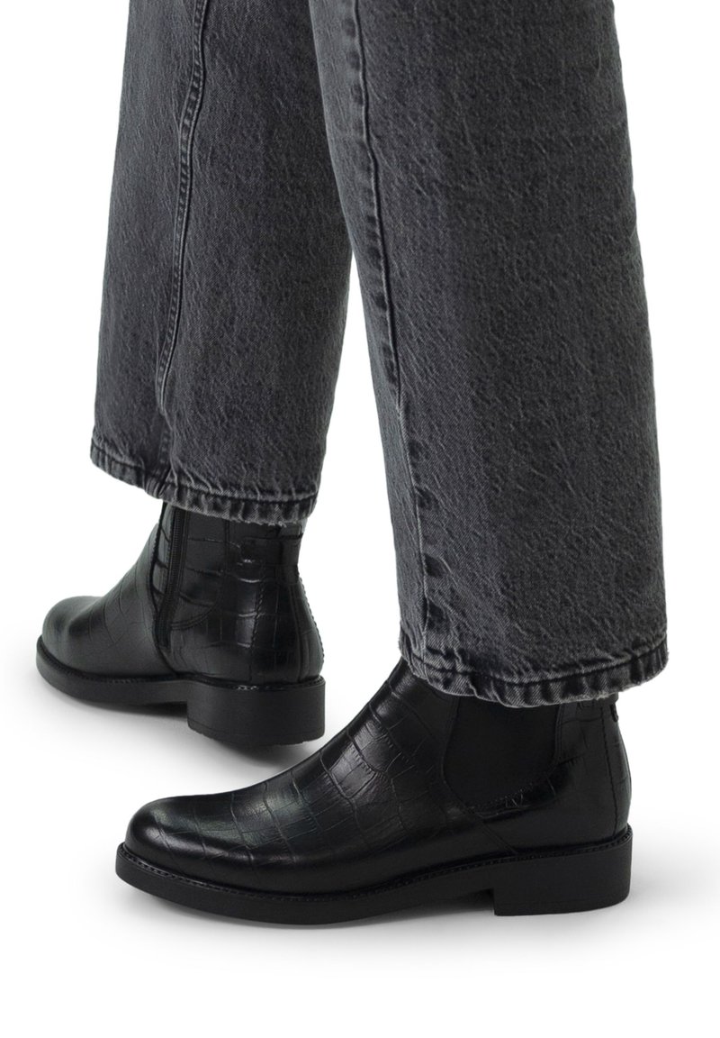 Stivaletti neri in pelle con texture a coccodrillo, dotati di pannelli laterali elasticizzati e un tacco basso e robusto, abbinati a jeans grigi a gamba larga.