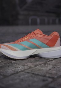 Lekki koralowo-turkusowy sportowy but do biegania na szarej kamiennej nawierzchni, widok z boku z widoczną białą podeszwą i sznurowadłami.