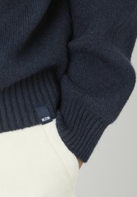 Pull en maille bleu marine avec des poignets et un ourlet côtelés, doté d'une petite étiquette noire portant la mention "BZE". Texture douce et coupe ajustée.