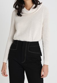 Cremefarbener Strickpullover mit einem Wasserfallausschnitt, kombiniert mit hoch taillierter schwarzer Hose mit kontrastierenden Nähten und Seitentaschen.