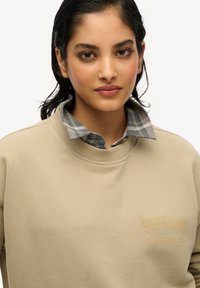 Sudadera beige con cuello redondo acanalado. Cuenta con un cuello de cuadros en azul y gris debajo. Texto dorado con tachuelas "Superdry Saint Tropez".