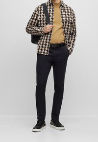 Muž v černých teniskách, černých kalhotách, béžové košili a černé a béžové vzorované bundě, nese černou tašku přes rameno na jednolitém pozadí.