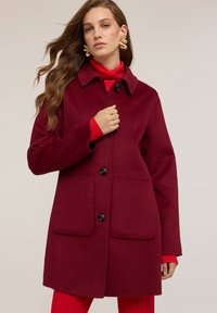 Cappotto in misto lana color bordeaux con collo a camicia, quattro bottoni frontali e due tasche a toppa. Tessuto liscio e design di lunghezza medio.