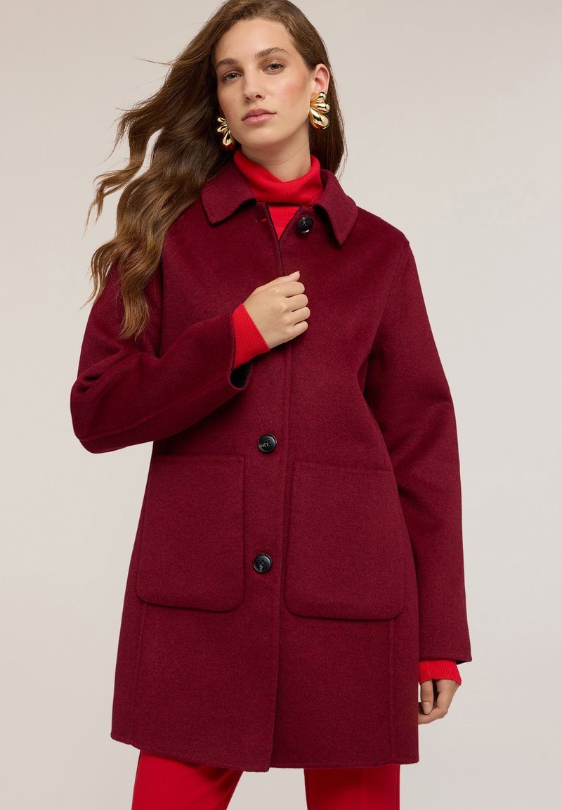 Cappotto in misto lana color bordeaux con collo a camicia, quattro bottoni frontali e due tasche a toppa. Tessuto liscio e design di lunghezza medio.