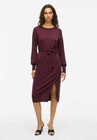 Burgundi spalvos megzta suknelė su ilgomis rankovėmis, dirželiu juosmens ir šoniniu įpjovimu. Lygus tekst�ūra ir prisiūtas siluetas, derinama su juodais atvirais kulnais.