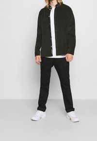 Mand iført sort skjorte over hvid T-shirt, sorte bukser og hvide sneakers står op ad en grå baggrund.