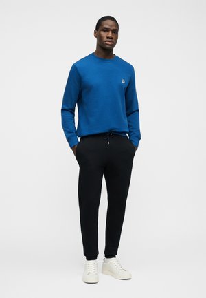 Man standing with hands in pockets wearing a blue sweatshirt, black jogger pants, and white sneakers against a plain background.