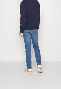 Man som bär blå jeans, beige och vita sneakers, samt en mörk marinblå sweatshirt, stående mot en helt vit bakgrund.