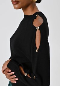 Donna che indossa un maglione nero con ritagli sulle spalle e sulle braccia, fissati da bottoni dorati, che mostra le sue mani piegate e parte del suo viso.