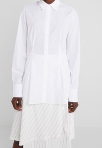 Chemise blanche à manches longues avec un col classique et une fermeture boutonnée à l'avant, présentant une texture lisse et une coupe droite. Portée sur une jupe rayée.