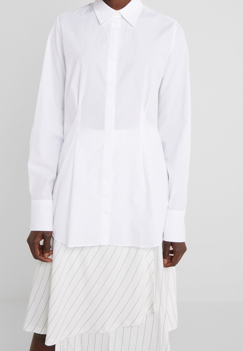 Chemise blanche à manches longues avec un col classique et une fermeture boutonnée à l'avant, présentant une texture lisse et une coupe droite. Portée sur une jupe rayée.