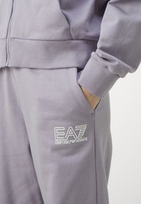 Svetra lila barve z rebrastimi manšetinami, z zadrgo in stranskimi žepi, okrašena z belim vezenim logotipom EA7 na prsih.