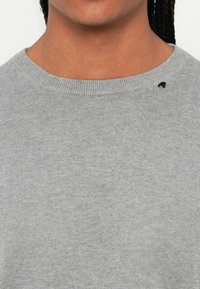 Personne portant un pull gris clair à col rond avec un petit logo noir brodé près de l'épaule droite.
