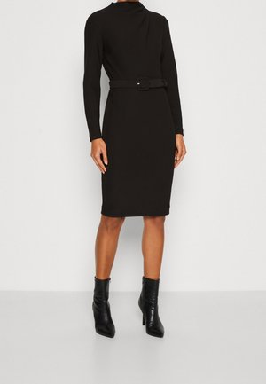 Kobieta stojąca w czarnej, długorękawowej sukience do kolan z paskiem oraz czarnych botkach z ostrym noskiem na białym tle.