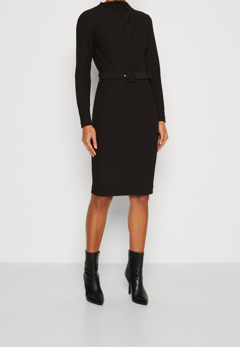 Femme debout portant une robe noire ceinturée à manches longues, longueur genou, et des bottines noires à bouts pointus sur fond blanc.