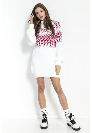 Kobieta w białej dzianinowej sukience z czerwonym wzorem, białe skarpetki z czerwonymi gwiazdkami oraz białe sznurowane botki z czarnymi podeszwami, stojąca przy białej ścianie.
