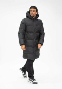 Doudoune noire avec capuche, au design long, sections matelassées horizontales et poches latérales. Portée sur un pantalon noir et des baskets.